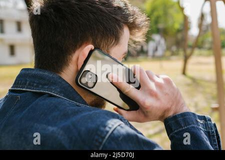 Junger Mann in blauer Jeans-Jacke von hinten mit Handy. Stehen Sie in einem Stadtpark, während Sie telefonieren. Verborgenes Gesicht mit Handy-Talki Stockfoto