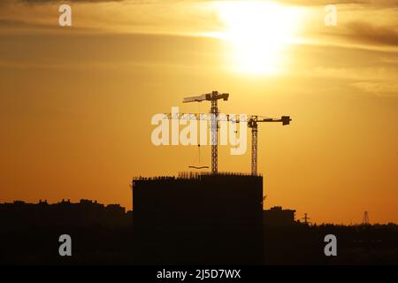 Silhouetten von zwei Turmdrehkranen und unfertigen Gebäuden bei Sonnenuntergang. Wohnungsbau, Wohnblock in der Stadt auf dramatischen Himmel Hintergrund Stockfoto