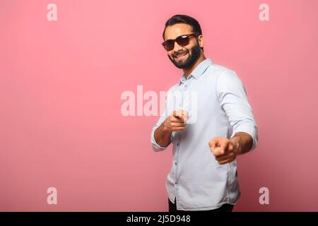 Gutaussehender lächelnder junger indischer Mann in Jeanshemd, der mit den Fingern auf Sie zeigt, isoliert auf rosa Hintergrund stehend, fröhlicher multirassischer Kerl, der auf die Kamera hinweist Stockfoto