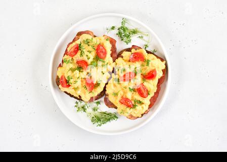 Rührei mit Microgreen, Speck und Tomaten auf Brot auf hellem Steingrund. Draufsicht, flach liegend Stockfoto