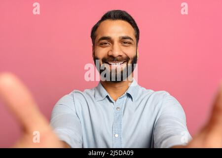 Porträt eines bärtigen, stattlichen indischen Mannes, der Selfie macht, die Kamera aus der Perspektive betrachtet und einen multirassischen, braunhaarigen Kerl, der sich selbst aufzeichnet. Innenaufnahme des Studios isoliert auf rosa Hintergrund Stockfoto