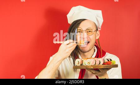 Junger Mann in Gläsern, als Koch gekleidet, hält Holzbrett mit Sushi-Rollen auf rotem Hintergrund. Männliche Köchin isst appetitliche Sushi-Rolle mit Essstäbchen Stockfoto