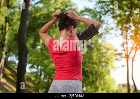 Rückansicht einer jungen, entschlossenen Sportlerin, die eine Smartphone-Halterung trägt, Pferdeschwanz bindet und sich auf die Flucht durch den Stadtpark im Freien vorbereitet Stockfoto