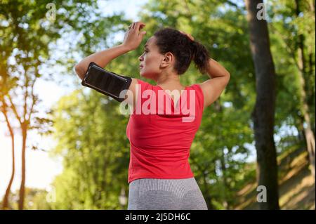 Rückansicht einer Sportlerin, die einen Smartphone-Halter trägt, Pferdeschwanz bindet und sich für den Lauf im Stadtpark im Freien vorbereitet Stockfoto