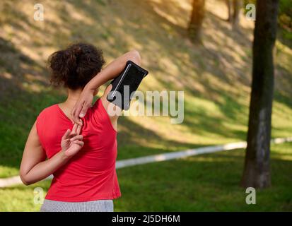 Rückansicht einer Afrikanerin, Sportlerin, die eine Smartphone-Halterung trägt, beim Training im Freien, beim Aufwärmen und Ausstrecken ihres Körpers während der m Stockfoto