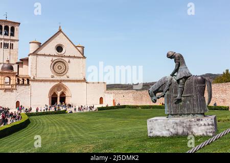 Basilika San Francesco d'Assisi mit Menschen. Vorderfassade der oberen Basilika mit grüner Wiese, Touristen auf Pilgerfahrt und Bronzestatue eines kn Stockfoto