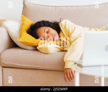 Glückliche und lächelnde afroamerikanische Frau, die sich auf der Couch niederlegt und Filme auf dem Laptop anschaut Stockfoto