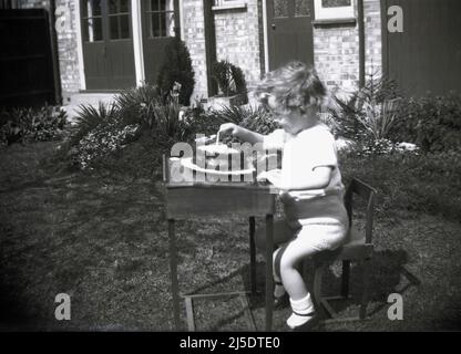 1950s, historisch, draußen in einem Garten, ein kleines Mädchen, das an einem alten hölzernen Schulschreibtisch sitzt und ihren Geburtstagskuchen anschaut, England, Großbritannien. Stockfoto