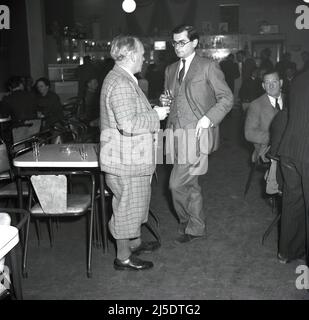 1950s, historisch, zwei gut gekleidete Männer, die mit einem Getränk in der Hand sprachen. Einer ist in einem Karo-Anzug mit plus Vieren, der andere in Anzug & Krawatte und Weste und sie stehen im Social Club der Steel Company of Wales im Abbey Stahlwerk in Port Talbot, Wales, Großbritannien, plaudern. Stockfoto