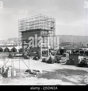 1950, historische Bauarbeiten beim Bau eines Betonwasserturms im riesigen Stahlwerk Abbey der Steel Company of Wales in Port Talbot, Wales, Großbritannien. Ein Schild auf der korragierten Hütte auf dem Boden in ihrer Nähe besagt: 'Temporäre Umspannstation'. Autos und Lieferwagen der damaligen Zeit stehen auf dem Parkplatz. Erbaut auf dem Gelände des alten Margam Eisen- und Stahlwerks, wurde das Abteiwerk 1951 eröffnet. Bis zum Jahr 1953, bis zum Jahr 1960s, war es das größte Stahlwerk in Europa. 1967 wurde das Unternehmen verstaatlicht und wurde Teil von British Steel. Stockfoto