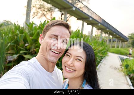 Ein multiethnisches Paar lacht während eines Selfies in einem Stadtpark während des Sonnenuntergangs Stockfoto