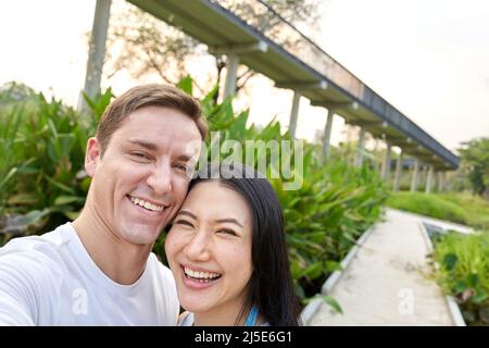 Ehepaar lacht zusammen, während sie ein Selfie in einem Park während des Sonnenuntergangs machen Stockfoto