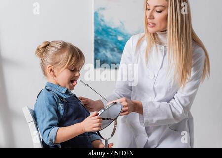 Logopädin in weißem Mantel mit logopädischer Sonde in der Nähe des Kindes mit Spiegel im Klassenzimmer Stockfoto