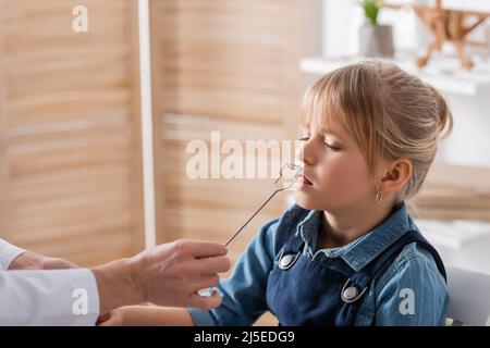 Logopädin im weißen Mantel hält Werkzeug in der Nähe Kind im Klassenzimmer Stockfoto