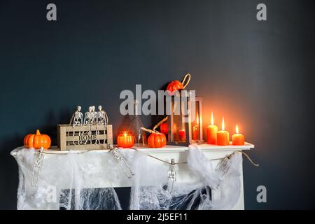 Halloween-Heimtextilien. Plastikspielzeug-Skelette in einer Holzkiste auf einem Kamin vor einer dunkelblauen Wand. Eine Girlande aus Skeletten. Spinnennetz auf dem Kleid Stockfoto