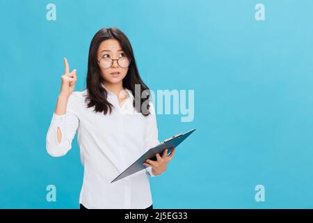 Aufgeregt süße asiatische Geschäftsfrau in klassischen Büro Kleiderordnung hält Ordner Tablette mit Checkliste zeigen Finger nach oben posiert isoliert auf blauem Studio Stockfoto