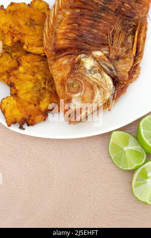 Gebratener Mojarra-Fisch, knusprig, traditionell im kolumbianischen Pazifik. Begleitet von Patakonen mit Haus und Zitrone, auf einem hellen Holzhintergrund.Copy Space. Stockfoto