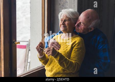 Senior Mann umarmt Senior Frau in der Nähe Fenster Senior Menschen unterstützen Konzept Raum drinnen Medium shot . Hochwertige Fotos Stockfoto