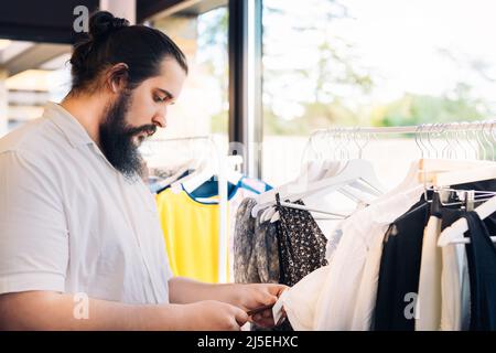 Junger Mann mit Bart, sucht und kauft Kleidung, im Modehaus. Einkaufskonzept. Stockfoto