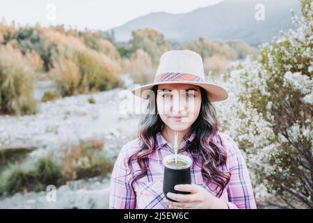 Junge kaukasische Frau mit Hut und blassrosa Bluse, trinkende Partnerin in einem natürlichen Raum mit herbstlicher Stimmung. Stockfoto