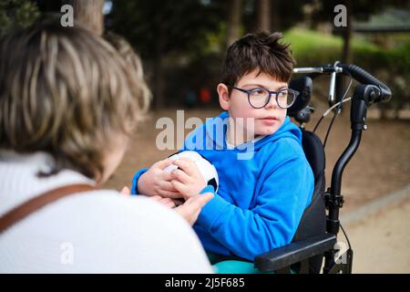 Behinderter Junge im Rollstuhl mit einem Fußball, während er mit seiner Mutter im Park spielt. Stockfoto