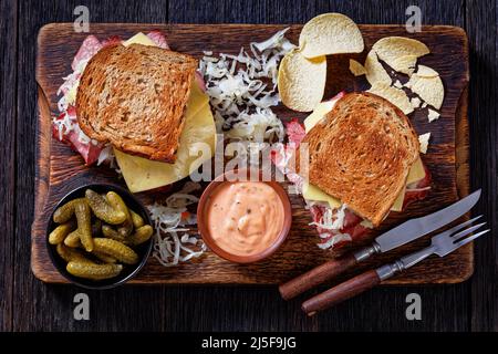 reuben-Sandwich mit Roggenbrot in dünne Scheiben geschnittenes Corned Beef, Sauerkraut, russischem Dressing und geschmolzenem Käse auf rustikalem Brett mit Gurken und Kartoffelchii Stockfoto