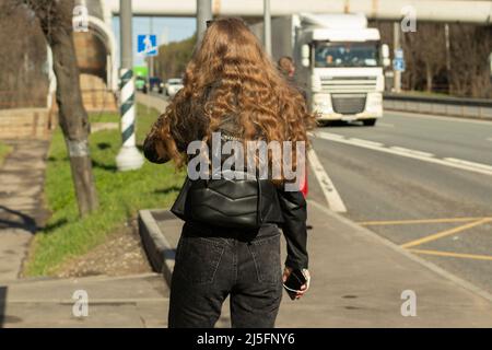 Mädchen wartet auf Bus auf der Straße. Frau mit lockigen Haaren. Schönes Mädchen schaut auf den Fluss der Autos. Warten auf ein Taxi in der Stadt. Student in schwarzer Kleidung. Stockfoto