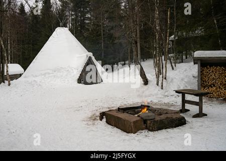 Rovaniemi, Finnland - 18.. März 2022: Ein Feuer im Freien zum Kochen neben einem schneebedeckten traditionellen samischen Holzhaus, auf finnisch Kota genannt. Stockfoto