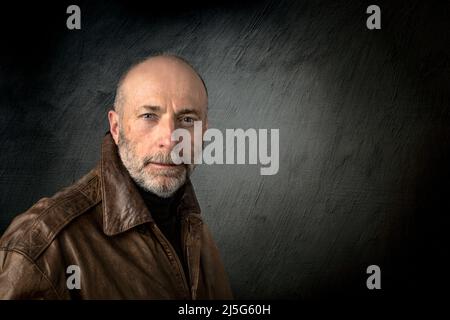 Selbstbewusster, grunzender Senior-Mann in Lederjacke – ein authentischer Kopfschuss vor dunklem Grunge-Hintergrund Stockfoto