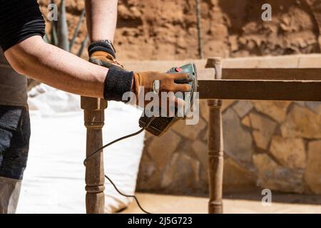 Mann, der mit Elektroschleifer an der Restaurierung alter Möbel arbeitet. Konzept für Holzpflege und Abdichtung Stockfoto