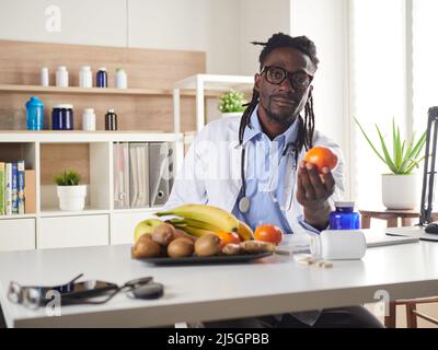 Der afroamerikanische Ernährungsberater schaut sich die Kamera an und zeigt gesunde Früchte in der Konsultation. Stockfoto