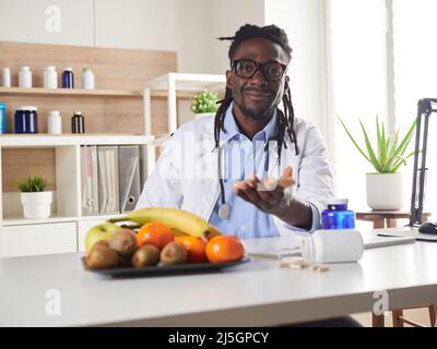 Der afroamerikanische Ernährungsberater schaut sich die Kamera an und zeigt gesunde Früchte in der Konsultation. Stockfoto