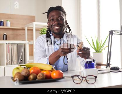 Der afroamerikanische Ernährungsberater schaut sich die Kamera an und zeigt gesunde Früchte in der Konsultation. Stockfoto