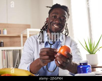 Der afroamerikanische Ernährungsberater schaut sich die Kamera an und zeigt gesunde Früchte in der Konsultation. Stockfoto