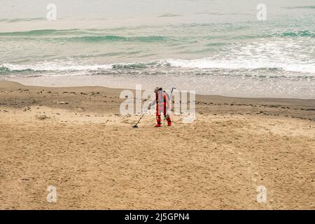 Ein Mann mit einem Metalldetektor geht in einem roten Anzug am Strand entlang. Zeitlupe, 4K, Zeitlupe Stockfoto