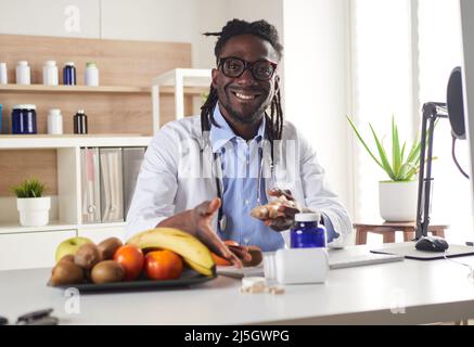 Der afroamerikanische Ernährungsberater schaut sich die Kamera an und zeigt gesunde Früchte in der Konsultation. Stockfoto