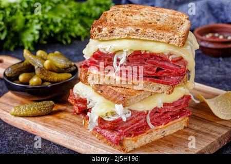 reuben-Sandwich mit Roggenbrot in dünne Scheiben geschnittenes Corned Beef, Sauerkraut, tausend Inseln Dressing und geschmolzener Käse an Bord mit Gurken und Kartoffellauch Stockfoto