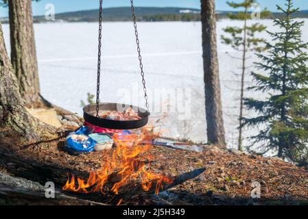 Bratpfanne voller Speck, der über einem Lagerfeuer hängt, gefrorener See und blauer Himmel im Hintergrund. Bild aus Vasternorrland Schweden. Fokus auf Vordergrund. Stockfoto