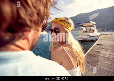 Attraktive, fröhliche Blondine flirtet mit ihrem Freund. Pärchen sitzt auf einem hölzernen Steg am Wasser. Zweisamkeit, Lifestyle, Luxuskonzept. Stockfoto