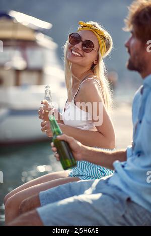 Lächelnde Frau, die auf dem Pier am See sitzt, in der Natur genießt und Bier trinkt Stockfoto