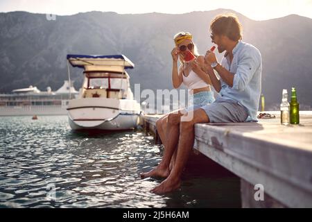 Liebend lächelndes Paar, das am Pier am See sitzt, die Natur genießt und Wassermelone isst Stockfoto