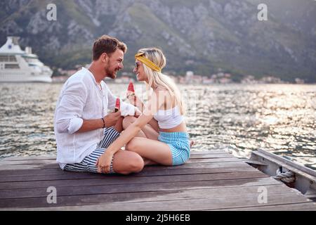 Romantischer Urlaub. Pärchen sitzt zusammen am Seeufer und genießt es Wassermelone zu essen Stockfoto