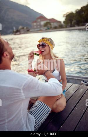 Romantischer Urlaub. Mann und Frau sitzen zusammen am Ufer des Sees und genießen es, Wassermelone zu essen Stockfoto