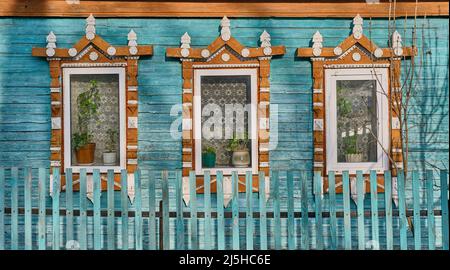 Fenster auf der türkisfarbenen Dielenwand des alten Hauses mit geschnitzten hölzernen Architraven mit rissiger Farbe. Stockfoto