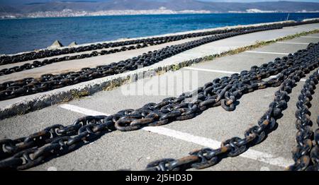 Große lange Seeketten aus rostig altem dunkelgrauem Metall für Schiffe und Boote liegen auf einem breiten steinernen leeren Pier, vor dem Hintergrund eines trüben blauen sp Stockfoto