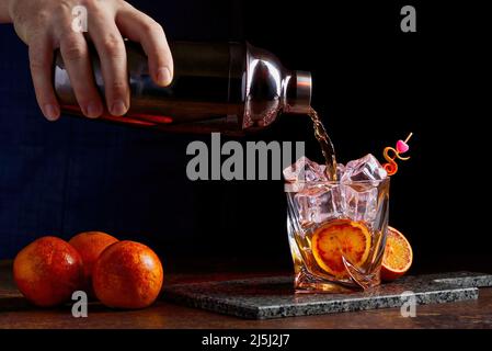 Alkoholcocktail, der aus dem Shaker in gless mit Orangenschalen-Eis auf dem Schreibtisch aus Stein gegossen wird. Stockfoto