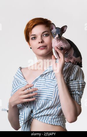 Sphynx Kätzchen sitzt auf der Schulter der schönen rothaarigen jungen Frau 25 Jahre alt. Weibliche Portraitfigur mit kurzen Haaren in gestreiftem weiß-blauem Hemd Stockfoto