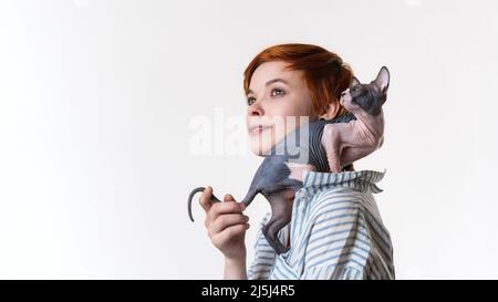 Lächelnde, rothaarige junge Frau mit der kanadischen Sphynx Cat auf ihrer Schulter. Porträt eines Hipsters in gestreiftem weiß-blauem Hemd. Studioaufnahme Stockfoto