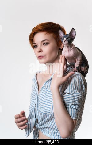 Sphynx Hairless Cat sitzt auf der Schulter der Rothaarigen jungen Frau. Hipster-Hündin mit kurzen Haaren und gestreiftem weiß-blauem Hemd. Studioaufnahme Stockfoto