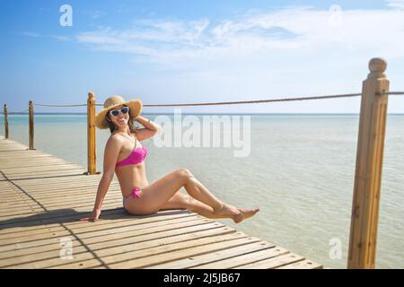 Glückliche junge Frau im Strohhut auf Holzponton sitzend Stockfoto